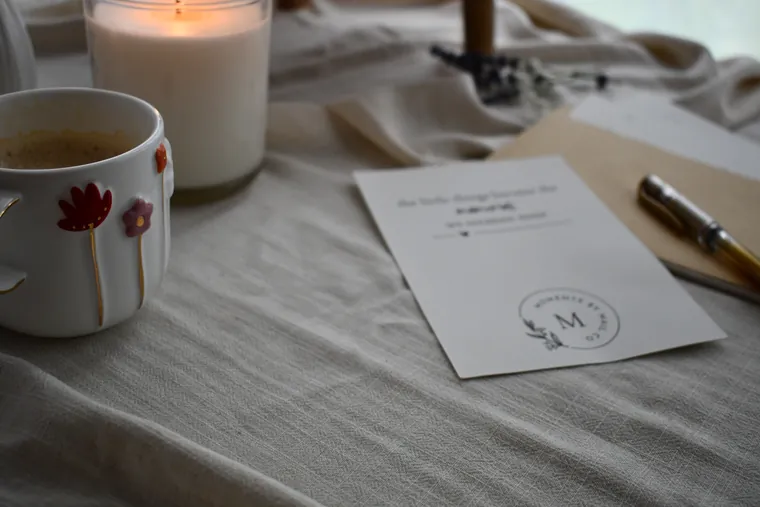 A meaningful gift card, pen, coffee, and candle arranged on linen.